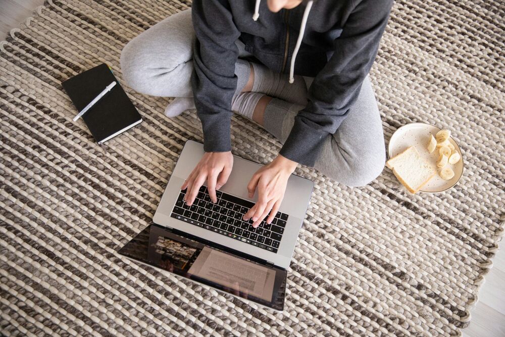 a person working on their laptop and eating while sitting on the carpet